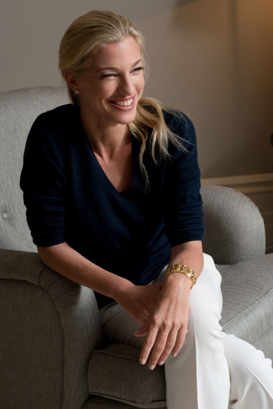 Nicole Junkerman seated on a pale sofa in a navy jumper, laughing in warm indoor light